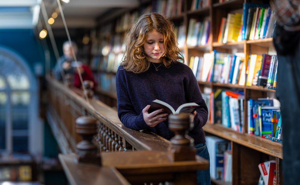 A teenage girl stands alongside a large bookcase reading a book.