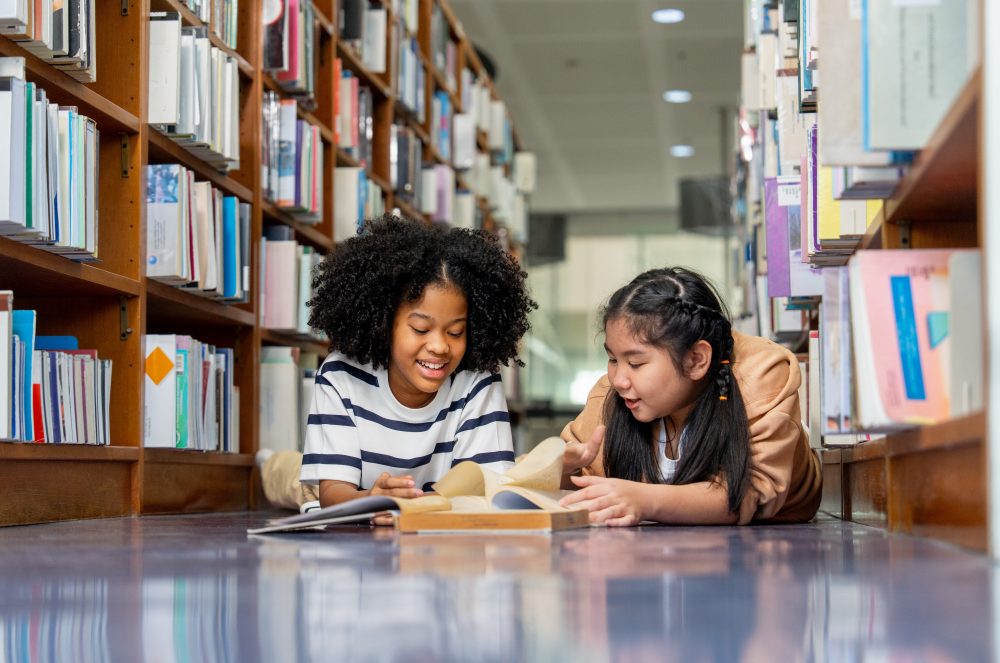 Two young girls lay on the floor of a library between shelves of books. They look together at an open book.
